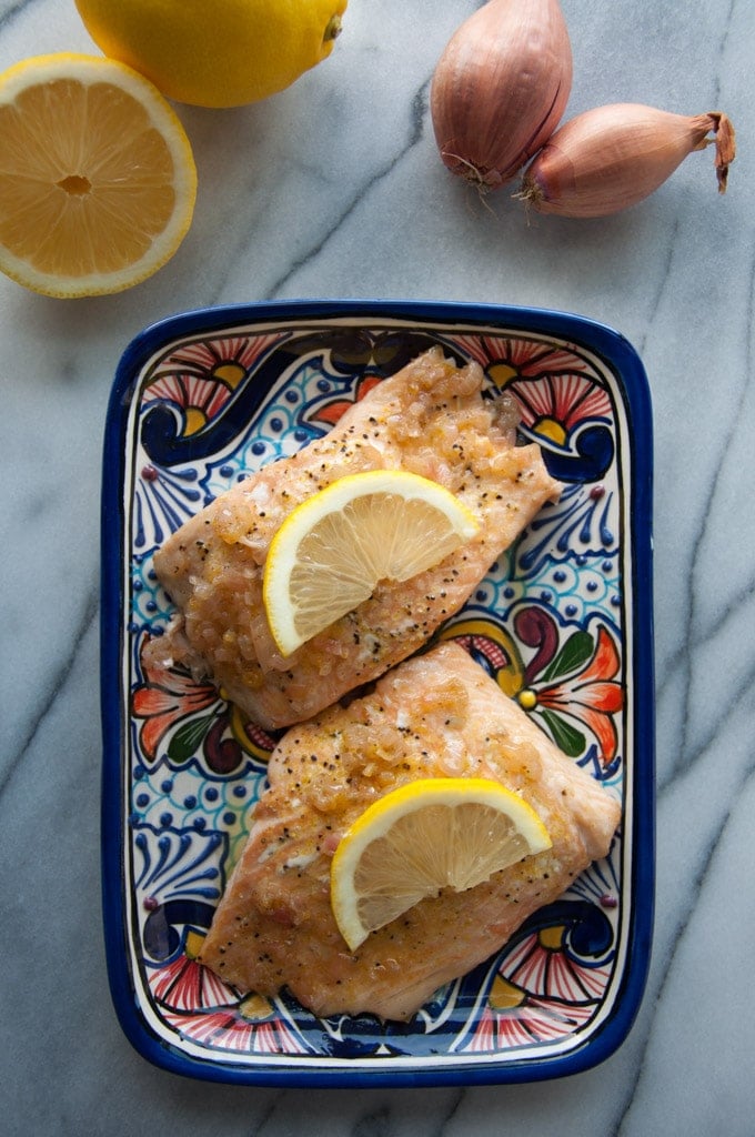 two pieces of shallot lemon butter salmon on a plate beside a lemon and two shallots