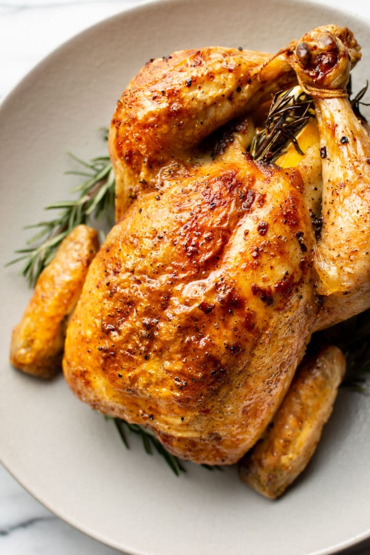 close-up of cooked crispy whole roast chicken in a shallow bowl surrounded by fresh rosemary