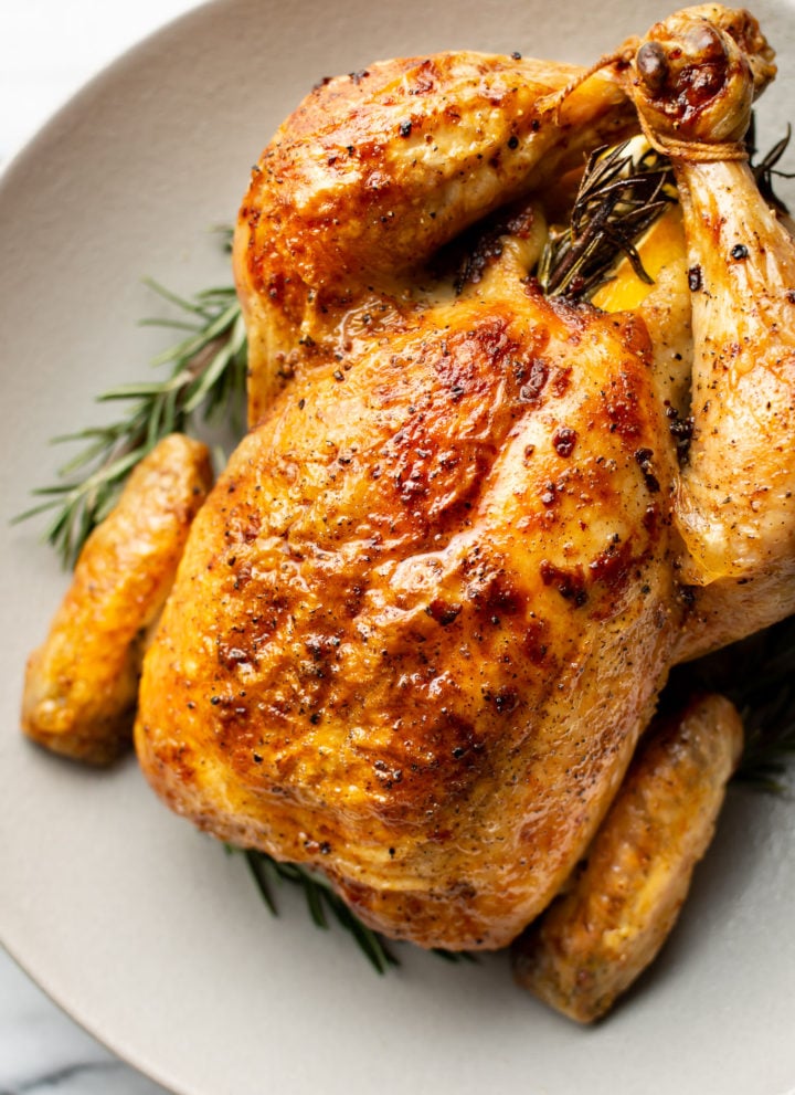 close-up of cooked crispy whole roast chicken in a shallow bowl surrounded by fresh rosemary