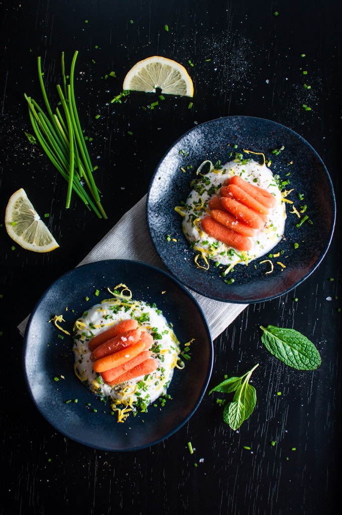 two black bowls of carrots with herb yogurt on a table with chives, mint, and a lemon slice