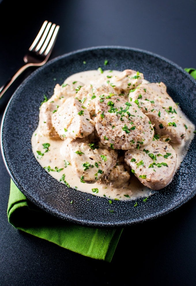 black plate with tender pork in a dijon sauce with parsley garnish beside a fork