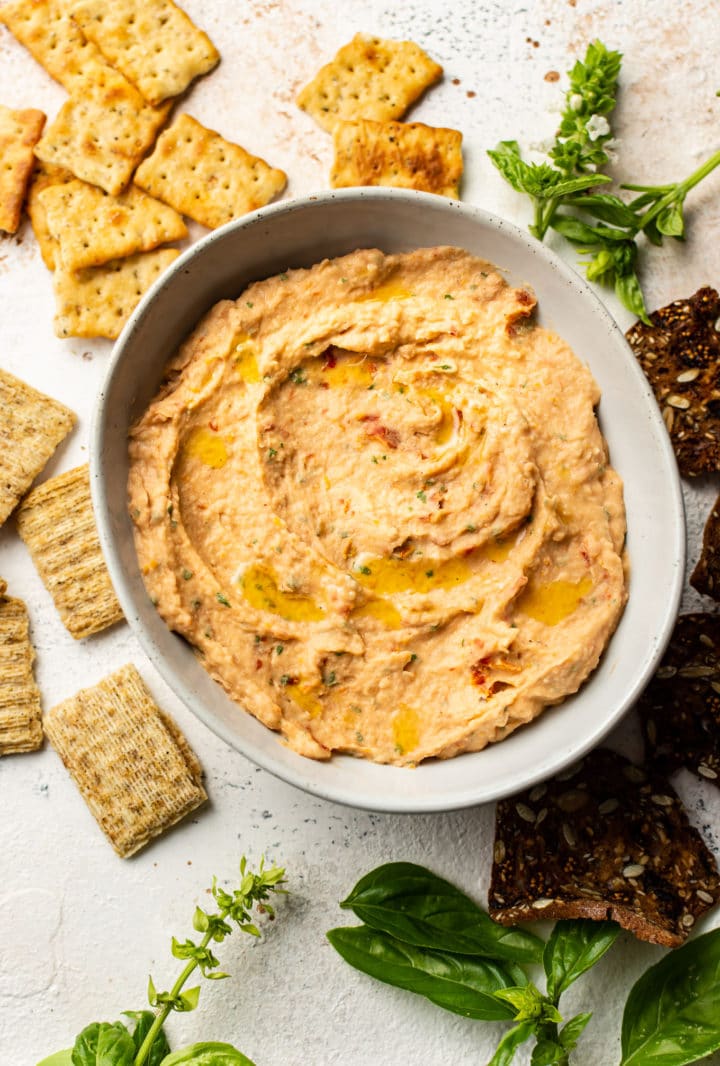 sun-dried tomato and basil white bean dip in a serving bowl, surrounded by crackers and fresh basil