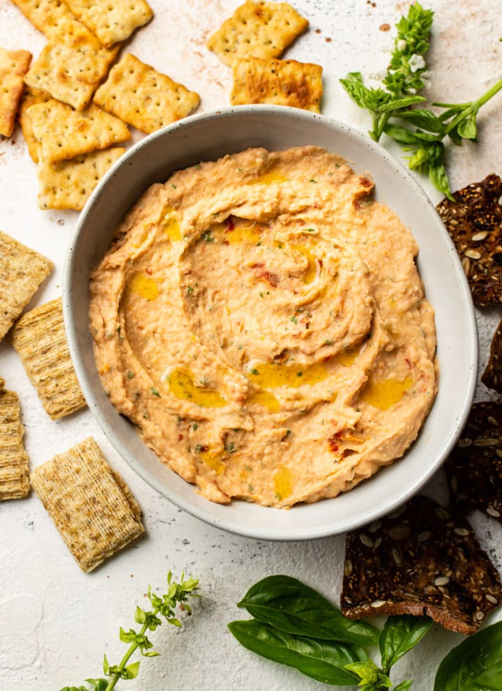 sun-dried tomato and basil white bean dip in a serving bowl, surrounded by crackers and fresh basil