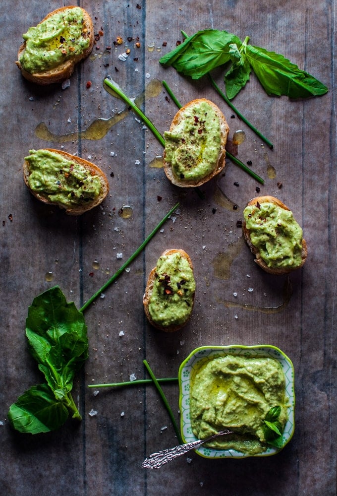 four slices of baguette topped with avocado pesto next to a bowl of avocado pesto with a spoon