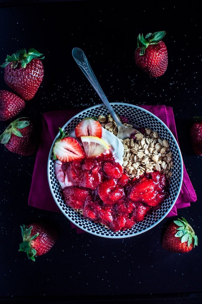 bowl of granola with Greek yogurt and strawberry vanilla lemon sauce with a spoon beside whole strawberries