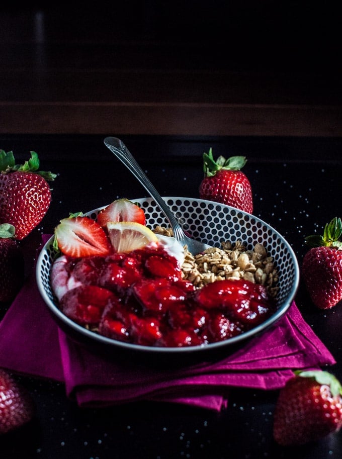bowl of granola with yogurt and a strawberry lemon vanilla sauce on a purple napkin beside whole strawberries