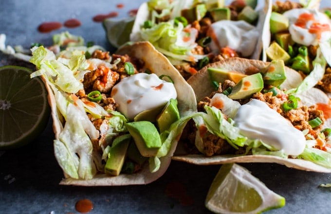 close-up of buffalo chicken tacos with hot wing sauce, avocado, sour cream, and lime wedges