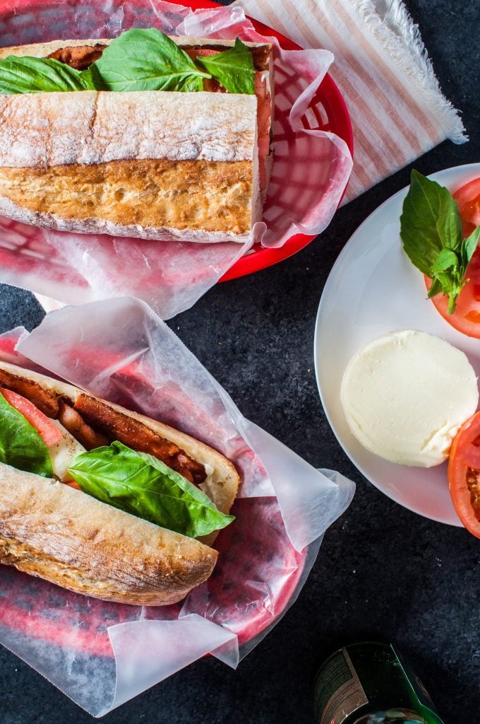 two red baskets with bacon Caprese sandwiches next to a plate of sliced tomatoes and mozzarella cheese