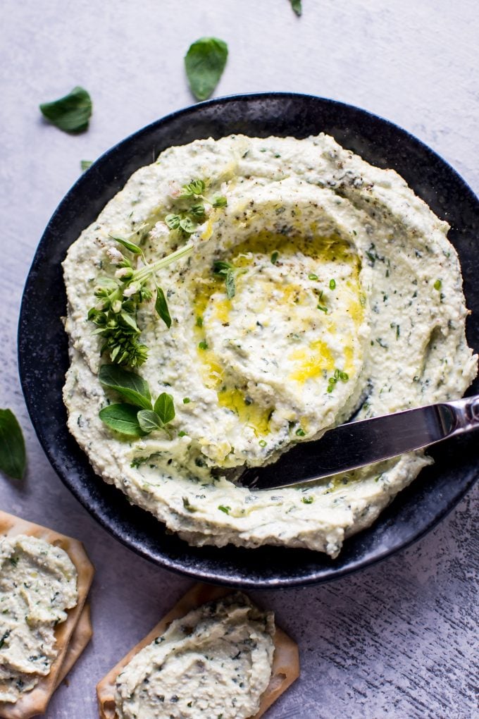 a butter knife in a bowl of whipped feta with fresh herbs and olive oil drizzle