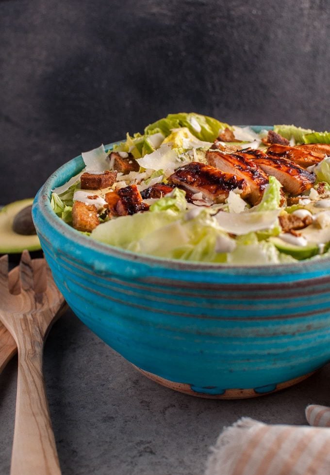 BBQ chicken Caesar salad in a large blue bowl beside wooden salad utensils