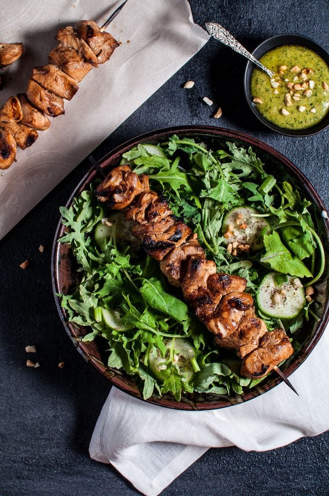 bowl of salad greens with a chicken kabob skewer on top next to small bowl of peanut lime dressing