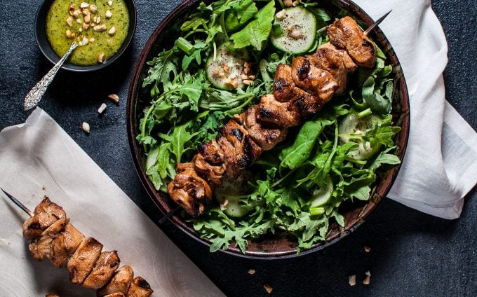 chicken kabob salad with peanut lime dressing in a bowl next to white cloth napkin, bowl of dressing, and chicken skewer