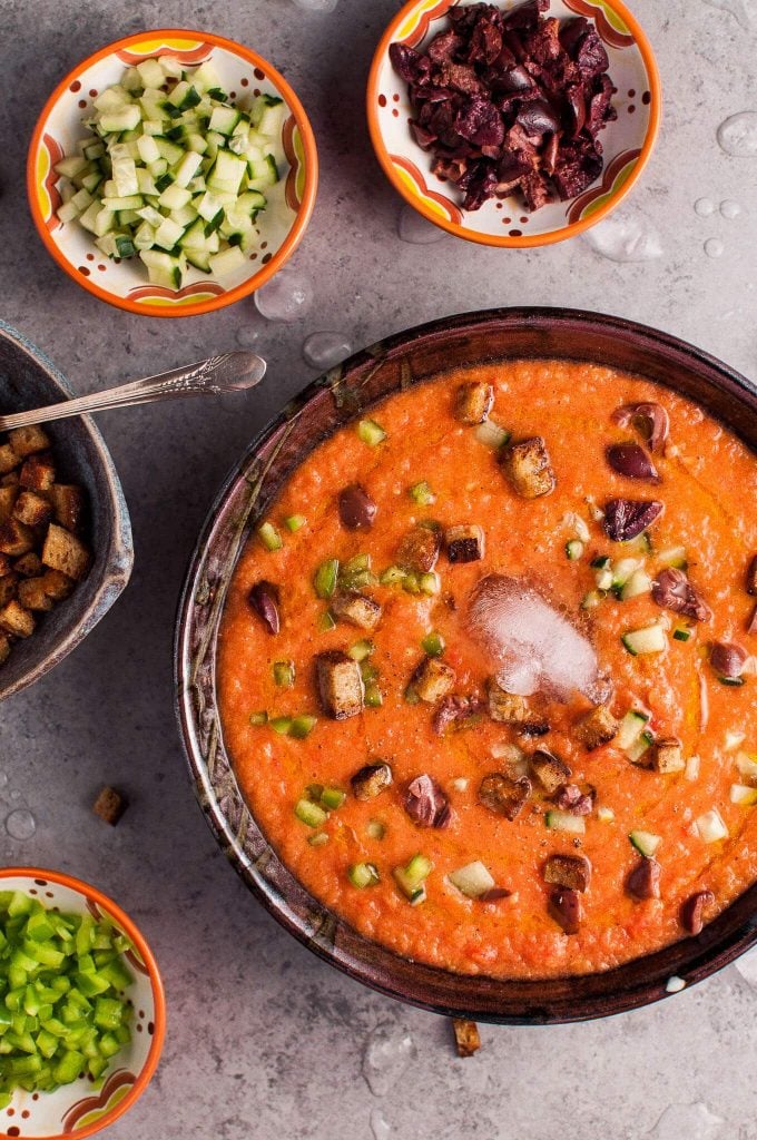 close-up of bowl with classic gazpacho soup with small bowls of croutons, olives, cucumber, and green pepper