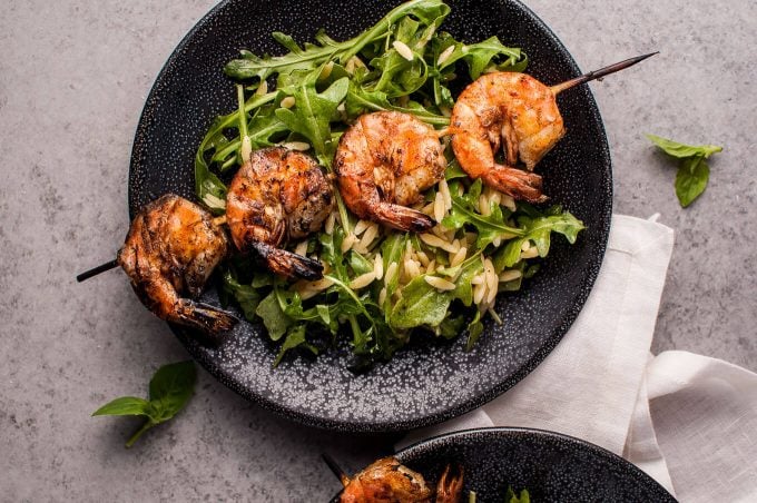black plate with a smoky grilled shrimp skewer, fresh arugula, and orzo
