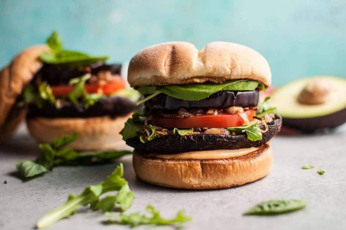 two grilled eggplant and mushroom burgers in buns beside half an avocado