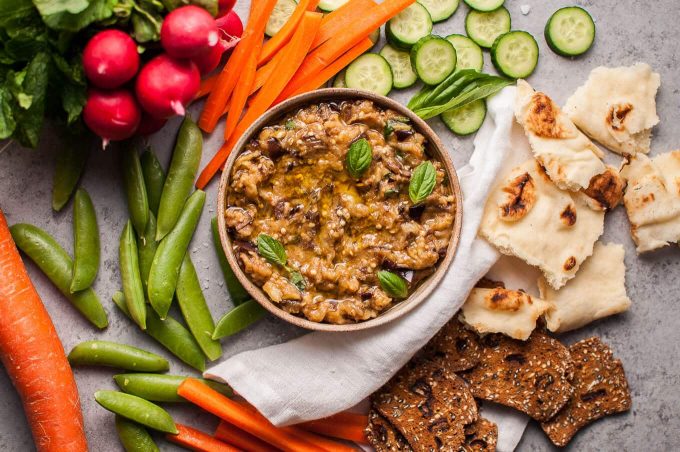 appetizer bowl of roasted eggplant dip next to crackers, pita bread, raw radishes, cucumber, carrots, and snap peas