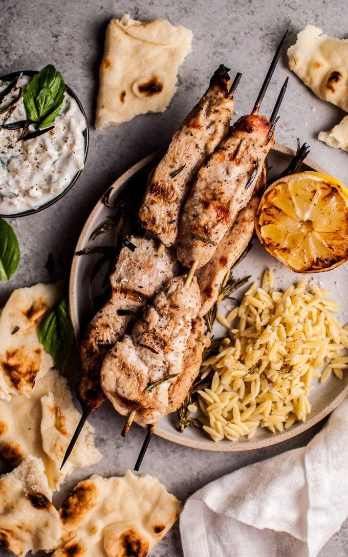 plate of rosemary chicken skewers, orzo, and lemon next to naan bread and small bowl of yogurt