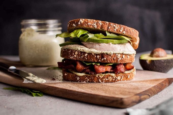 homemade green goddess chicken sandwich on a cutting board with bacon, avocado, and salad greens