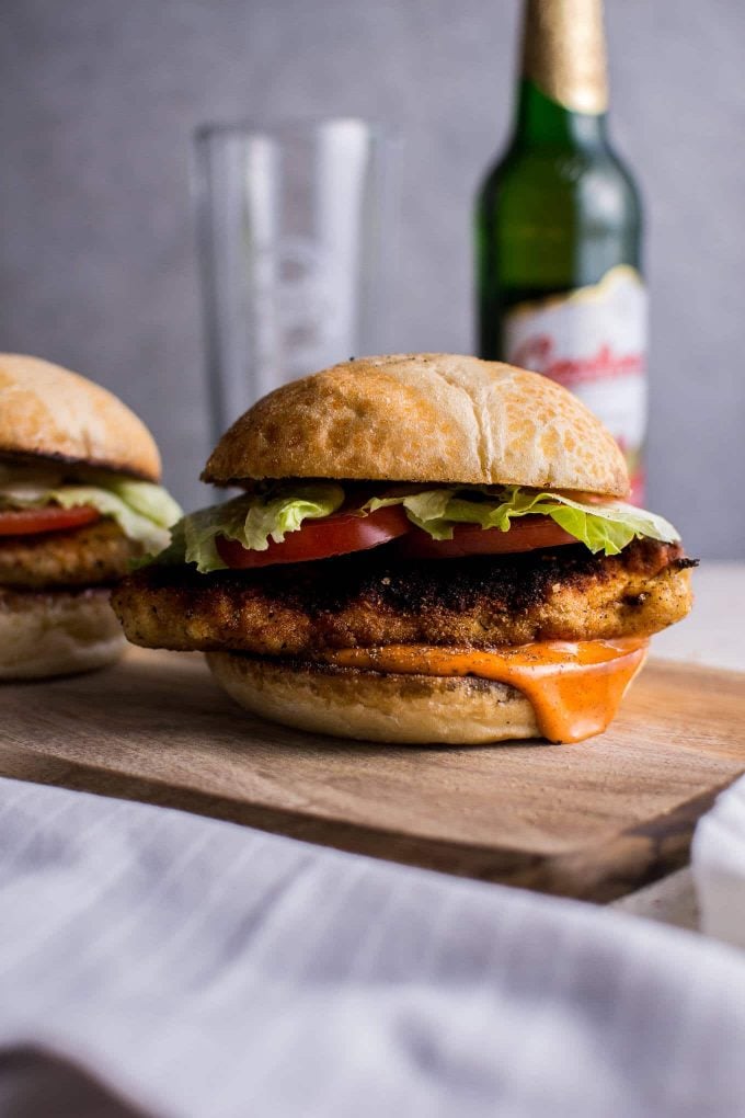 two spicy fried chicken sandwiches on a wooden surface with a bottle of beer and a glass