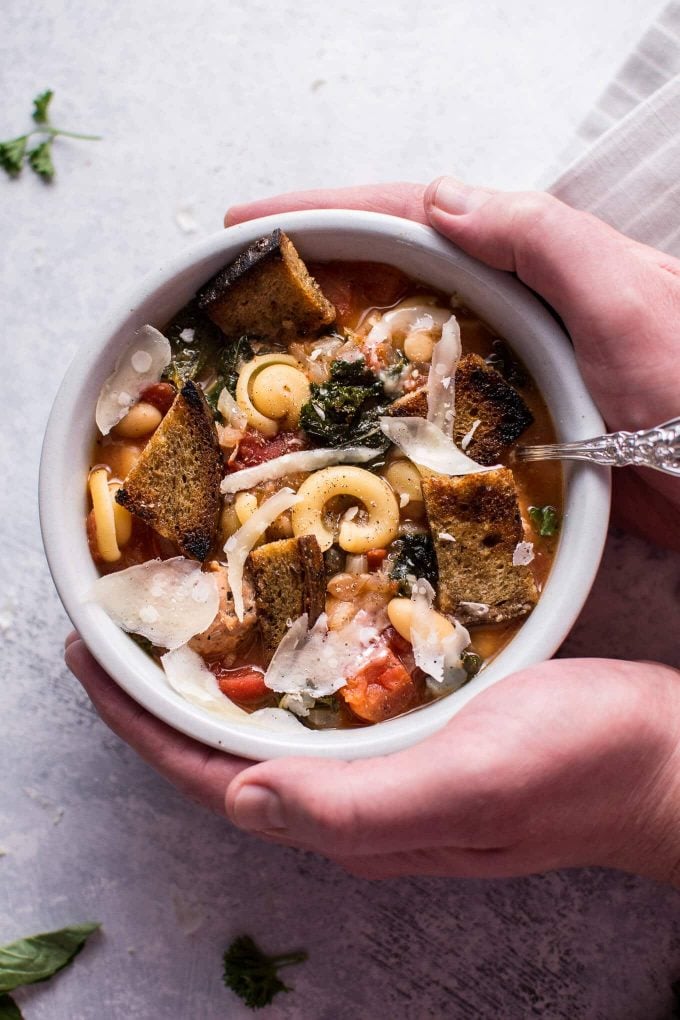 two hands holding bowl of tuscan chicken soup with pasta and kale