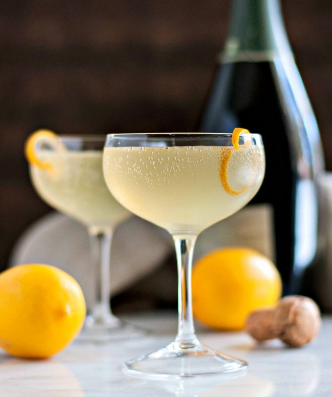 two cocktail glasses with meyer lemon french 75 cocktails with whole lemons, cork, and champagne bottle in background