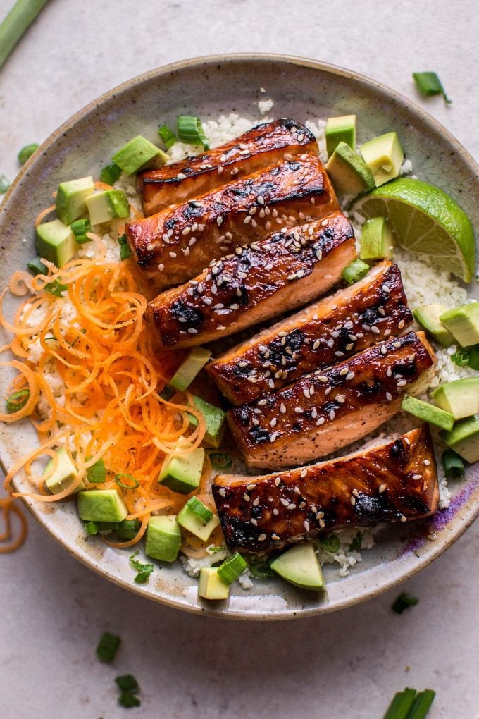 close-up of glazed hoisin and sesame salmon in a bowl with chopped avocado, carrots, cauliflower rice, and lime wedge