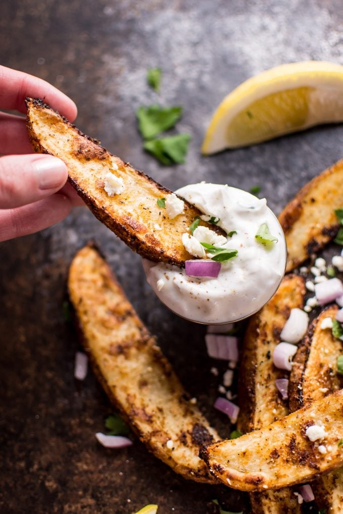 female hand dipping Greek loaded baked potato wedge in tzatziki dipping sauce