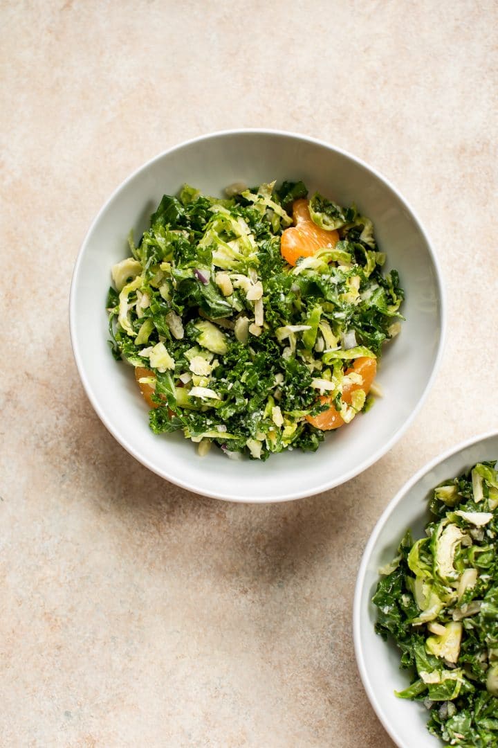a white bowl with shredded kale and Brussels sprouts salad with mandarin oranges, pecorino cheese, slivered almonds, red onions, and a honey mustard dressing