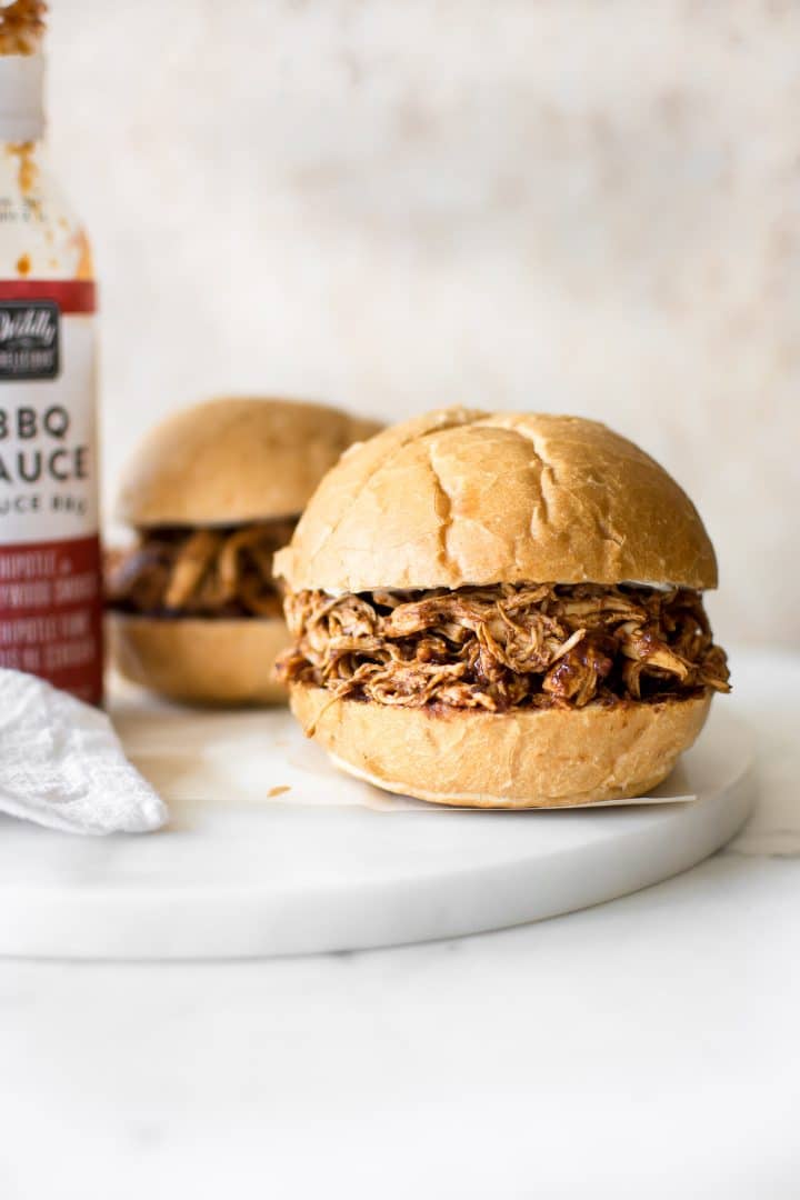 two Instant Pot pulled chicken sandwiches beside a bottle of barbecue sauce