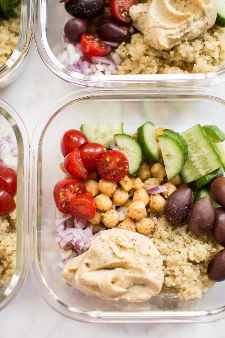 close-up of glass container with cucumber, olives, tomatoes, chickpeas, hummus, and quinoa