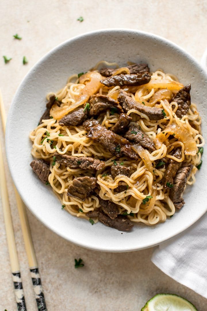 beef and ramen noodles on a white plate beside wooden chopsticks