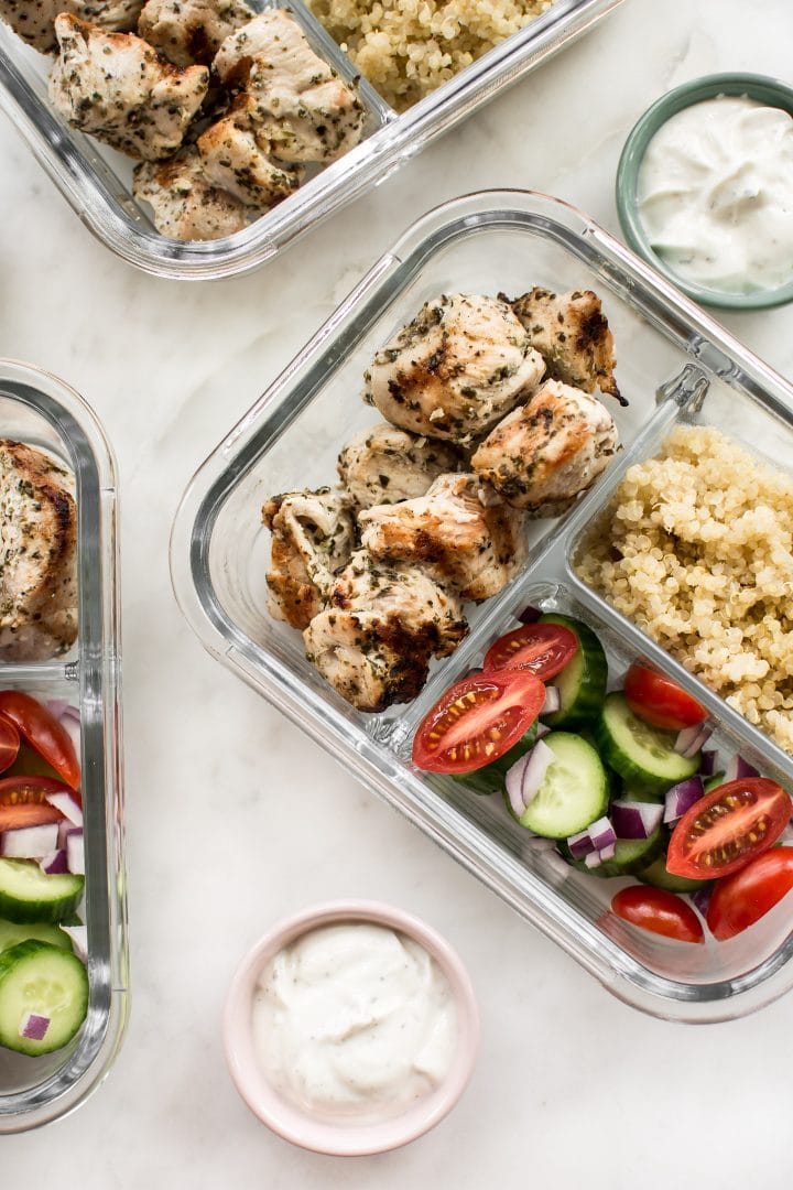 glass containers with grilled chicken, quinoa, tomatoes, cucumbers, and tzatziki in small bowls