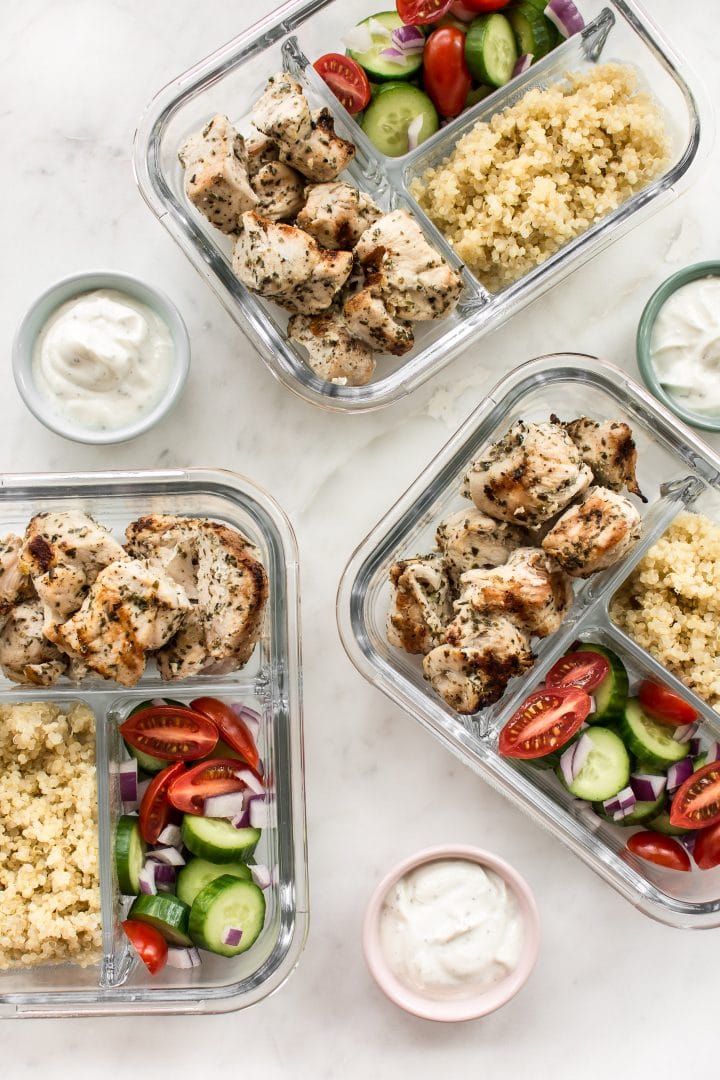 three glass meal prep containers of Greek chicken souvlaki with small tzatziki dip bowls
