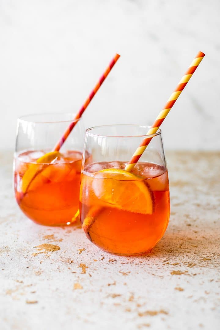 two aperol spritz cocktails in stemless glasses with straws and lemon slices