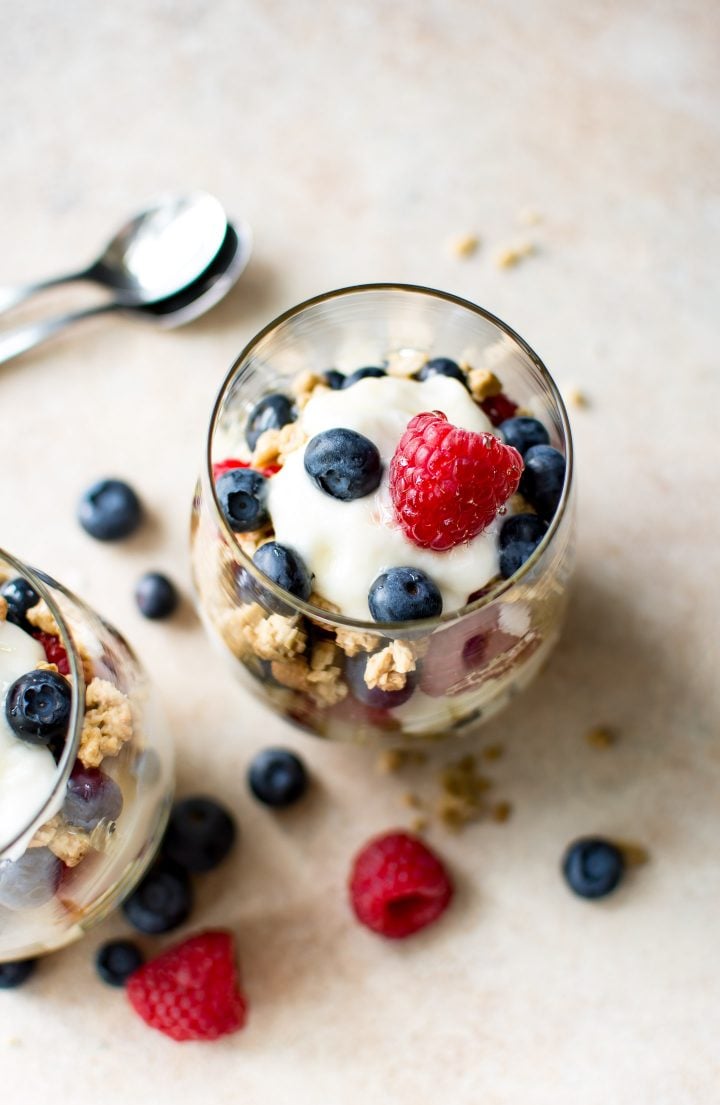 two berry parfaits on a table with two spoons and loose berries