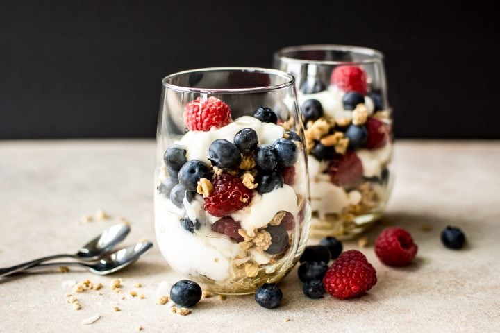 two clear glasses with blueberry and raspberry parfaits on a surface beside spoons
