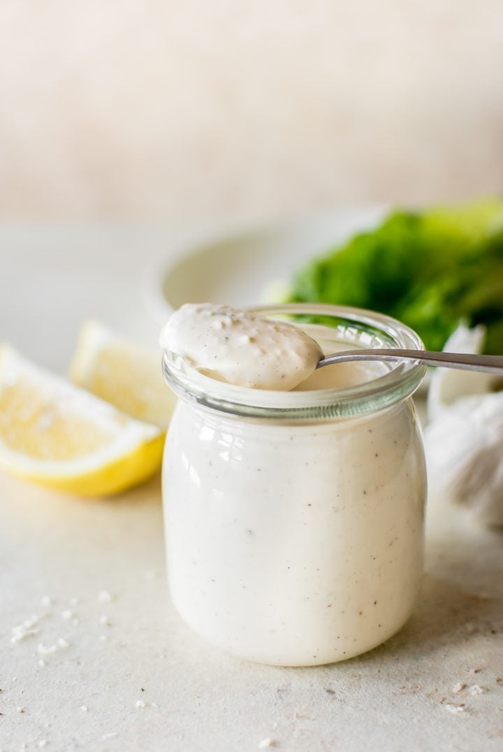 homemade caesar dressing in a clear glass yogurt jar with a spoon and lemon slices