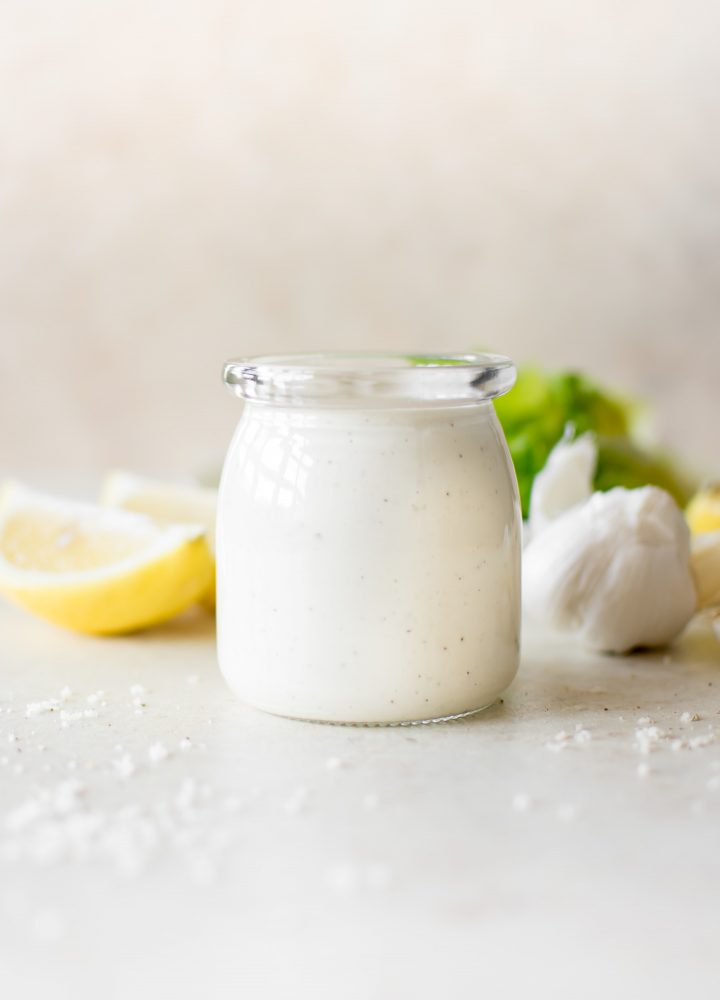 homemade caesar salad dressing in a clear glass yogurt jar beside lemon slices and a head of garlic