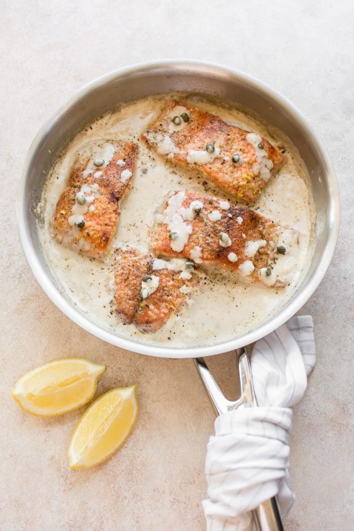 creamy salmon piccata in a metal skillet with a white tea towel and two lemon wedges