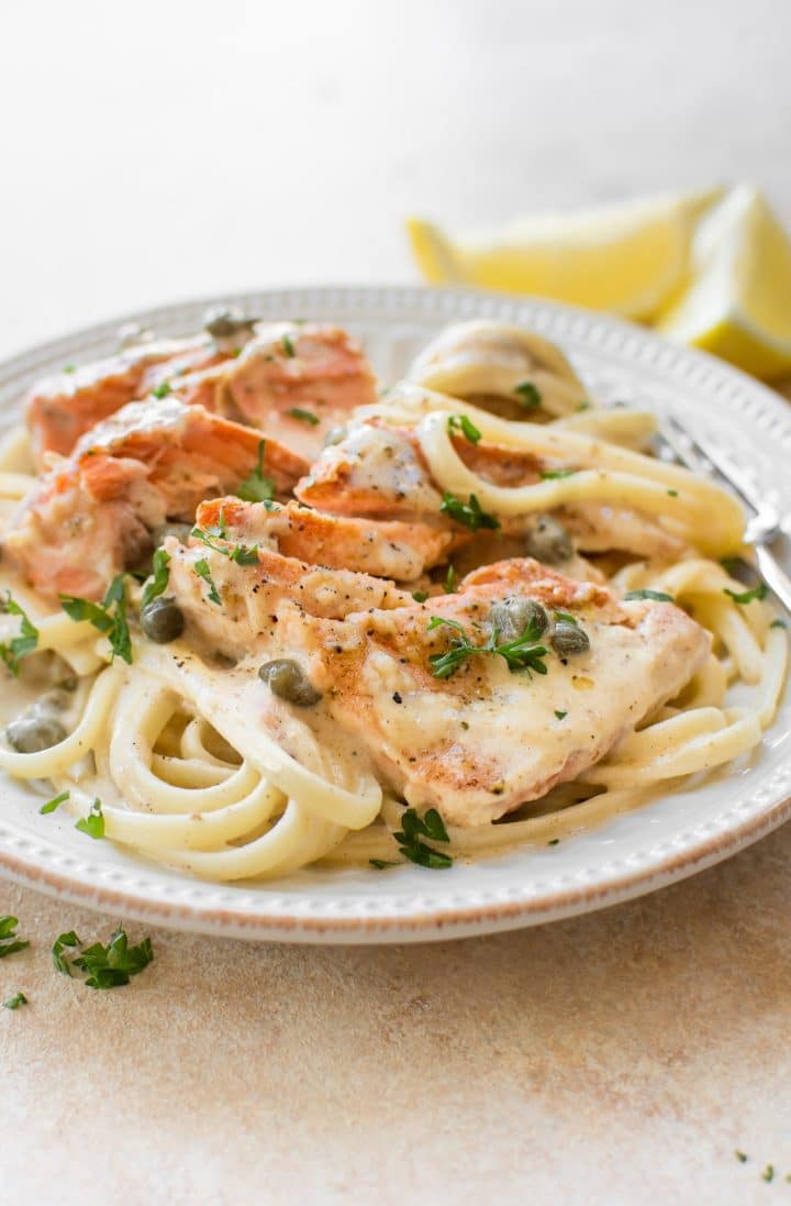 creamy salmon piccata over pasta on a white plate beside two lemon slices and a fork