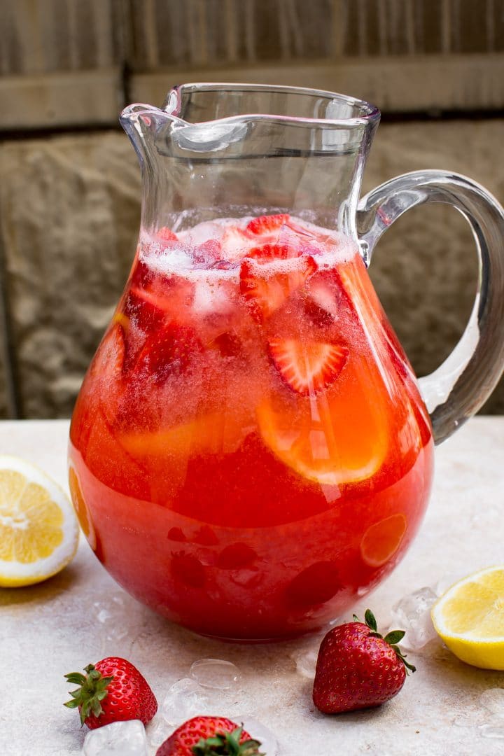 homemade strawberry lemonade on a table in a large glass pitcher beside lemon wedges