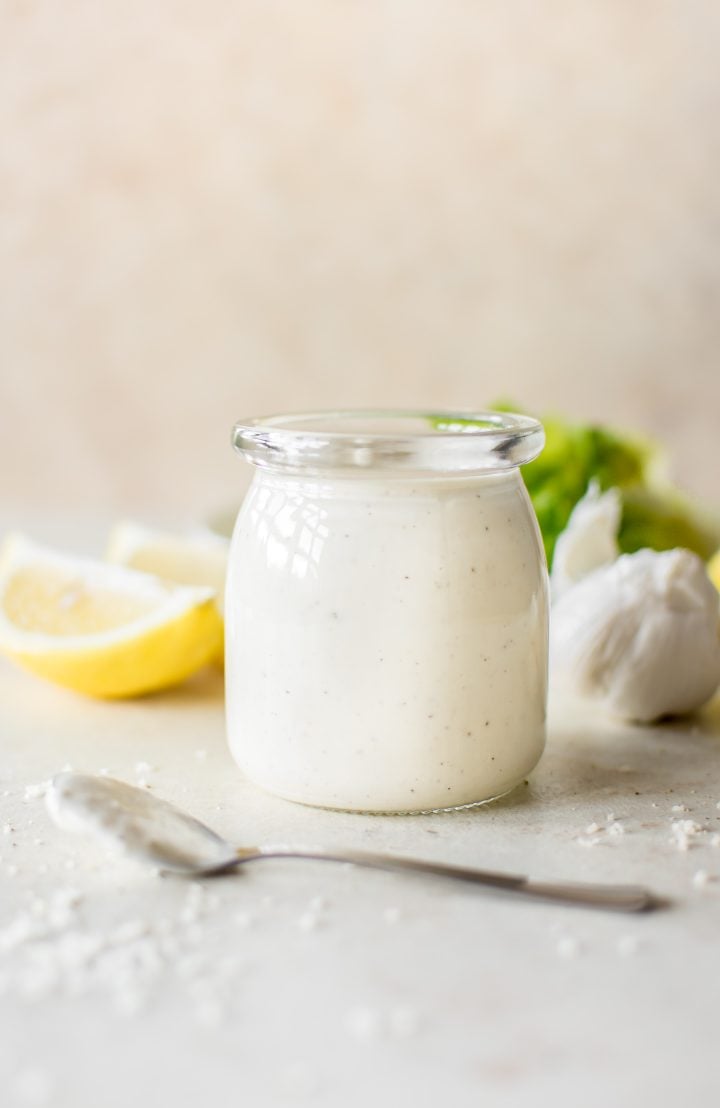 caesar salad dressing in a glass jar beside a spoon, head of garlic, and lemon slices