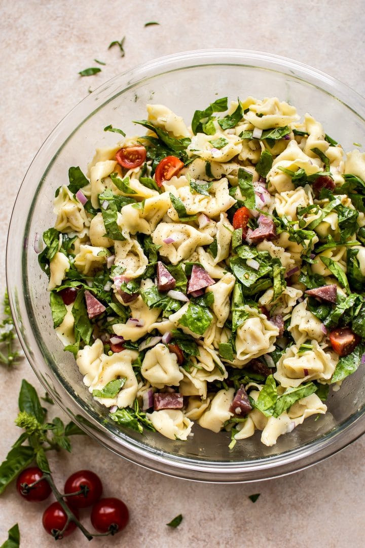 spinach tortellini salad with salami pieces in a clear glass bowl