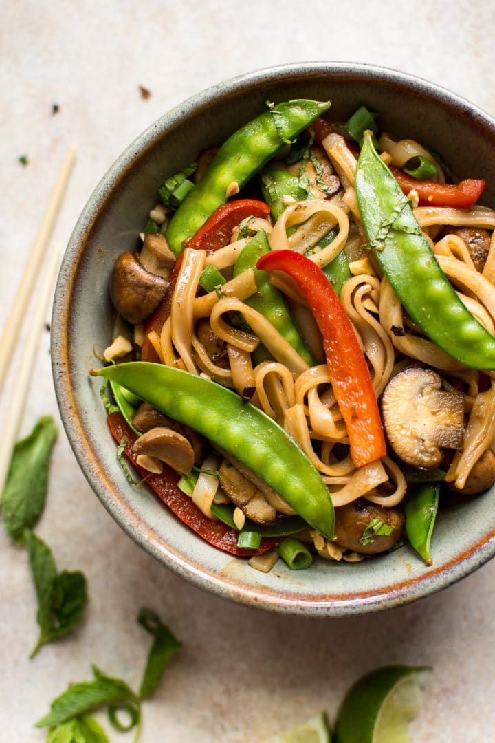 close-up of vegan rice noodle stir fry with peas, mushrooms, and red peppers in a bowl beside chopsticks