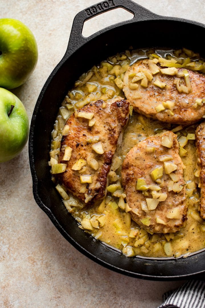 pork chops with chopped apples in a cast iron skillet beside two whole granny smith apples