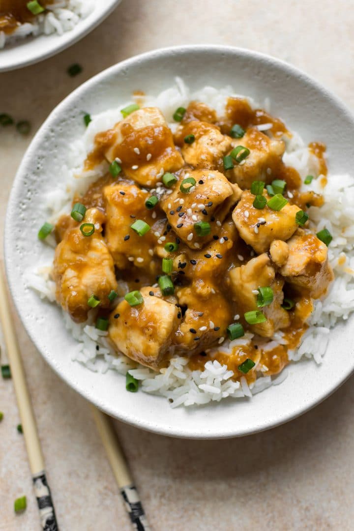 white bowl with tender mongolian chicken over rice topped with sesame seeds beside chopsticks