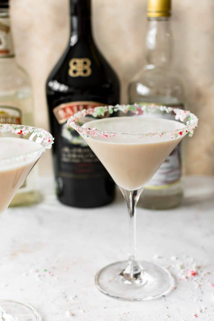 Christmas cocktail recipe with Baileys, Creme de Cacao, Peppermint Schnapps, and a crushed candy cane rim in a martini glass with bottles in the background