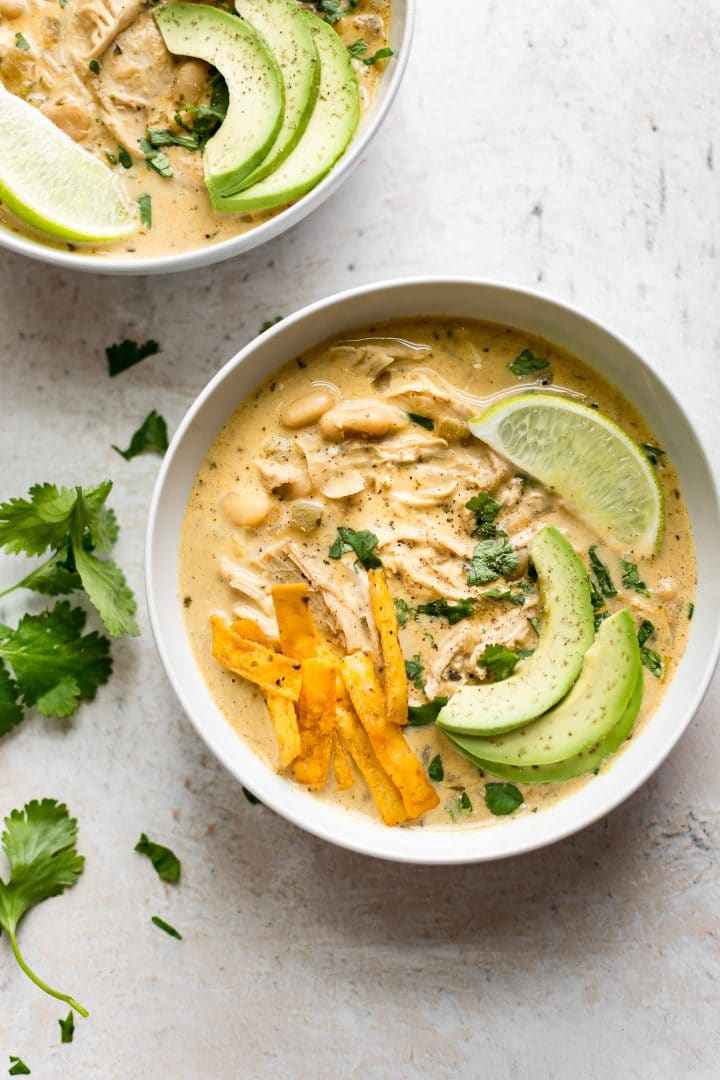 two white bowls of Instant Pot creamy white chicken chili topped with avocado, tortilla chips, and lime
