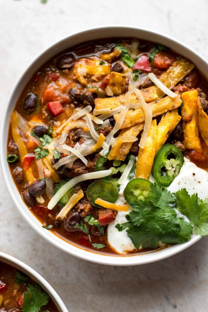 close-up of beef and black bean chili in a white bowl topped with cilantro, sour cream, and tortilla chips