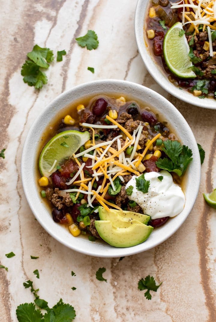 two white bowls of easy Crockpot taco soup with ground beef, corn, beans, avocado, lime, and cilantro